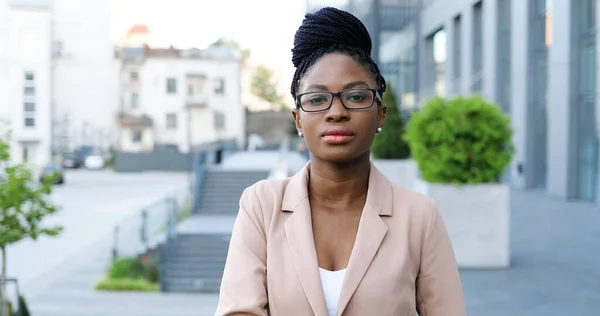 Portré felvétel fiatal afro-amerikai gyönyörű nő szemüvegben nézi a kamerát a szabadban, és támaszkodik az állát a kezét. Vonzó magabiztos, komoly üzletasszony az utcán, közel az üzleti központhoz. — Stock Fotó