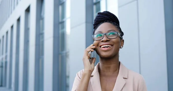 Afroamerikansk ung vacker glad kvinna i glasögon talar på mobiltelefon och ler utanför på businesscenter. Glad affärskvinna i glasögon som talar i mobiltelefon. Samtal. — Stockfoto