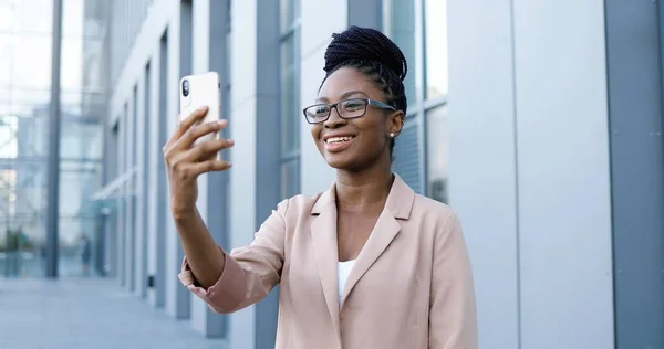 꽤 행복 한 아프리카계 미국인젊은 여성 안경을 끼고 비즈니스 빌딩의 스마트폰 야외에서 비디오 채팅을 하고 있습니다. 아름다운 여성이 휴대폰으로 웹캠을 통해 이야기하고 비디오를 보여 주는 모습. — 스톡 사진