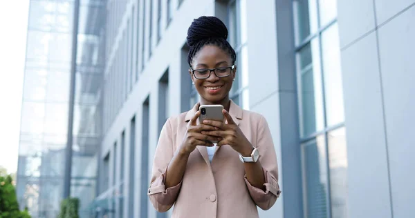 아프리카계 미국인 행복 한 젊은 사업가가 스마트폰으로 도청하고 검색 합니다. 여성이 밖에서 휴대 전화로 문자 메시지를 보내고 있습니다. 여자들은 대화를 나누면서 기기를 사용하고 미소를 짓는다. — 스톡 사진