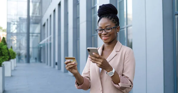 Afroamerikansk ung vacker kvinna i glasögon med smartphone, kreditkort och shopping på nätet. Vacker kvinnlig affärskvinna knacka på mobiltelefon och köpa varor på internet. Köpare på gatan. — Stockfoto