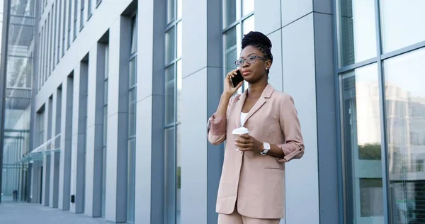 Afro-americano giovane bella donna in occhiali parlare al cellulare al di fuori del business center. Fiduciosa donna d'affari con gli occhiali che parla al cellulare e tiene in mano una tazza di caffè da asporto. — Foto Stock