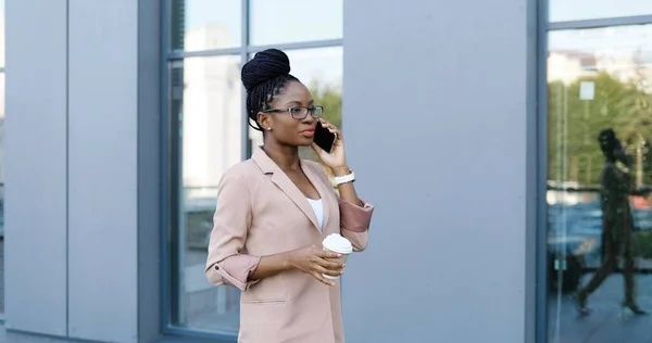 Mulher bonita afro-americana de óculos falando no celular, bebendo e andando fora. Empresária elegante falando no celular, bebendo café para ir de manhã e pisando. — Fotografia de Stock