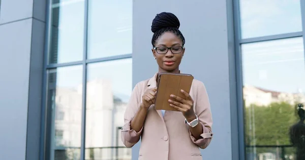 Afroamerikansk ung affärskvinna i glasögon som knackar och bläddrar på surfplattan. Kvinnans sms på datorn utomhus. Självsäker kvinna som använder gadget och chattar. — Stockfoto