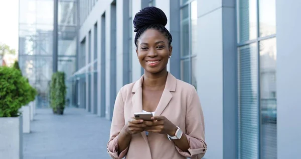 아프리카계 미국인젊은 사업가가 스마트폰으로 두드리고 검색하며 카메라 앞에서 웃는 모습. 여자가 밖에서 휴대 전화로 문자 메시지를 보내고 있어. 전자 기기를 사용하고 수다를 떨고 있는 여성. — 스톡 사진