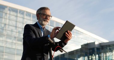 Yaşlı, gri saçlı, gözlüklü, bisikletin önünde duran ve tablet cihazını tıklatan ya da kaydıran bir adam. Yaşlı büyükbaba alet bilgisayarı kullanıyor ve açık havada internette geziniyor..