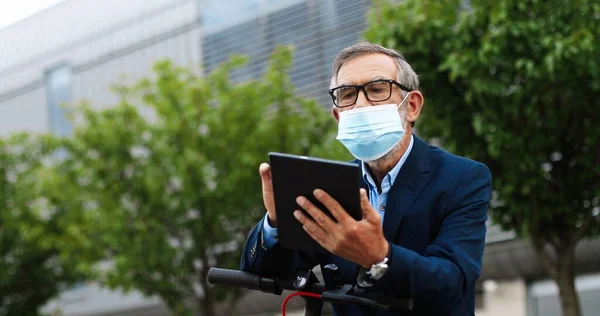 백인 노인은 안경과 의료용 마스크를 착용하고 자전거와 usinf 태블릿 장치에 앉아 회색 머리를 하고 있다. 밖에 있는 가젯 컴퓨터로 안경을 두드리고 스크롤을 하고 있는 나이든 남자. — 스톡 사진
