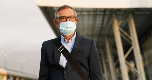 Retrato de hombre viejo caucásico guapo en gafas y máscara médica de pie al aire libre en el entorno urbano y mirando a la cámara. Hombre mayor de pelo gris en anteojos y protección respiratoria. —  Fotos de Stock