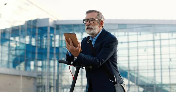 Senior muž v brýlích stojící na kole na ulici a klepání nebo rolování na tabletu zařízení ve velké skleněné moderní budově. Starý dědeček pomocí gadget počítač a prohlížení online venkovní. — Stock fotografie