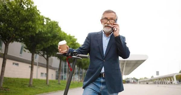 Starší muž v brýlích, stojící na elektrickém skútru ve městě, drží před sebou kávu a mluví na mobilu. Starý děda mluví venku na mobilu. Telefonát na ulici. Rozhovor. — Stock fotografie