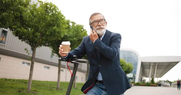 Starší muž v brýlích, stojící na elektrickém skútru ve městě, pije kávu a mluví na mobilu. Starý děda mluví na mobilu a popíjí horký nápoj. Telefonní hovor na ulici. — Stock fotografie