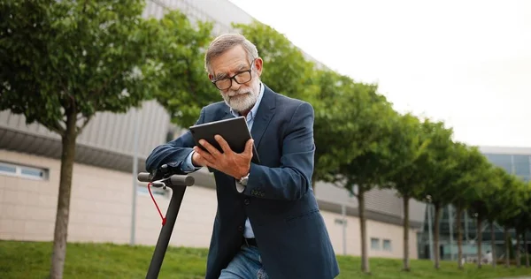 Senior muž v brýlích stojící na elektrickém skútru na ulici ve městě a klepání nebo rolování na tablet zařízení. Starý dědeček pomocí gadget počítače a prohlížení on-line venku. — Stock fotografie