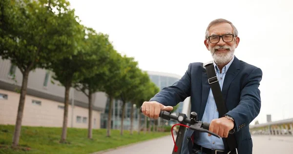 Retrato de homem velho caucasiano com cabelos grisalhos e em óculos sentados em bicicleta e olhando para a câmera. avô grisalho sênior em scooter elétrico ao ar livre. Homem aposentado em bicicleta na cidade. — Fotografia de Stock