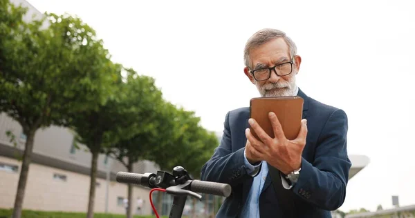 Starší šedovlasý muž v brýlích a sluchátkách, stojící na kole na ulici a poklepávající nebo posouvající se po počítači. Starý dědeček v brýlích pomocí tabletu zařízení a prohlížení online venkovní. — Stock fotografie