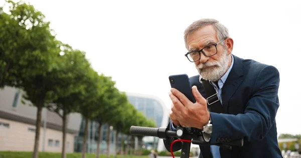 Starší šedovlasý muž v brýlích a sluchátkách, stojící na kole na ulici a poklepávající nebo posouvající se po mobilním telefonu. Starý dědeček v brýlích pomocí smartphone a prohlížení online venkovní. — Stock fotografie