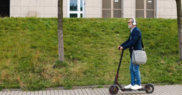 Caucasiano velho homem de cabelos grisalhos em óculos e fones de ouvido em pé na scooter elétrica e andando em algum lugar. reformados do sexo masculino ou empresário dirigindo veículo urbano e ouvindo música ao ar livre. — Fotografia de Stock