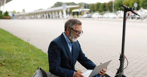 Starý běloch v brýlích sedí venku na zelené trávě u elektrického skútru a videochatuje na notebooku. Muž mluví přes webkameru na ulici. Dědeček má videochat a mává rukou. — Stock fotografie