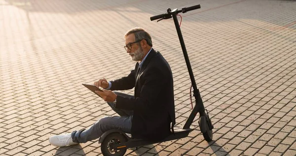 Grauhaariger alter Mann mit Brille sitzt auf einem Elektroroller auf der Straße und tippt oder blättert auf einem Gadget-Computer. Senior Opa nutzt Tablet-Gerät, schaut zu und surft online im Freien. — Stockfoto