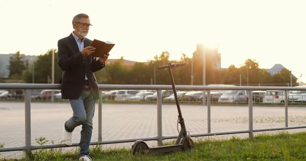 Kavkazský starý pohledný muž v brýlích, stojící venku, čtoucí knihu a naklánějící se plot. Starší dědeček četl učebnici nebo příručku. Vedle je elektrický skútr. Parkování aut. — Stock fotografie