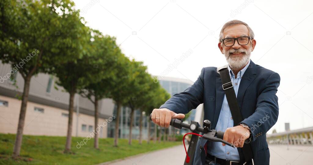 Portrait shot of Caucasian old man with gray hair and in glasses sitting on bike and looking at camera. Senior gray-haired grandfather on electric scooter outdoors. Male pensioner on bicycle in city.