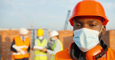 Portrait of African American handsome young man constructor in casque and medical mask standing outdoor at construction and looking at camera. Close up of male builder at building in helmet. Pandemic. clipart