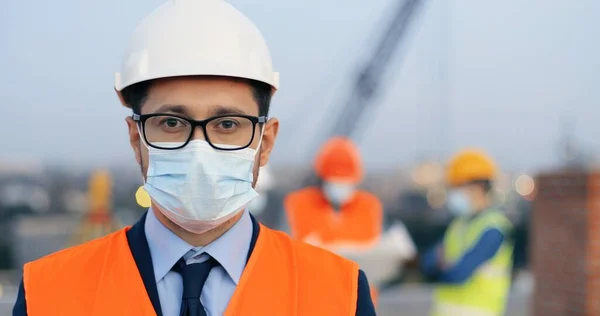 Portret białego, przystojnego, młodego konstruktora w trumnie, okularach i masce medycznej stojącego na zewnątrz przy budowie i patrzącego w kamerę. Zbliżenie inwestora biznesowego przy budowie kasku. — Zdjęcie stockowe