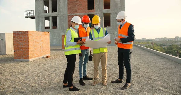 Team of mixed-races males and female constructors in helmets and medical masks standing at building top and discussing constructing plan draft. Pandemic concept. Engineers and architectors.