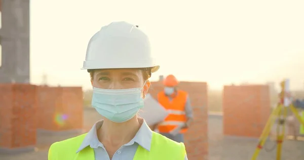 카스 크와 의료용 마스크를 착용하고 야외에 서 있는 백인 여성 건설자의 초상화. 헬멧을 쓰고 건물을 짓는 여성 건축가의 근접 사진입니다. 코로나 바이러스. 배경 작업을 하는 건축가들. — 스톡 사진