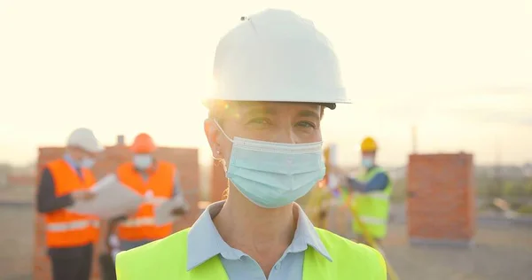 Portret Kaukaska młoda konstruktorka w kasku i maska medyczna stojąca na zewnątrz przy budowie. Zamykam żeński budowniczy w kask. Koronawirus. Budowniczowie pracujący w tle. — Zdjęcie stockowe