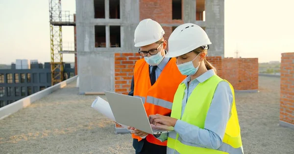 Casal caucasiano de construtores masculinos e femininos em capacetes e máscaras médicas conversando e discutindo esboço de plano no lado da construção. Homem e mulher construtores que trabalham na construção com computador portátil. — Fotografia de Stock