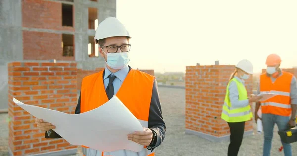 Kaukaski młody biznesmen inwestujący w hemet i maskę medyczną stojący przy wysokim budynku i patrzący na projekt budowy w rękach. Mężczyzna inżynier lub architekt na stronie konstrukcyjnej pandemii. — Zdjęcie stockowe