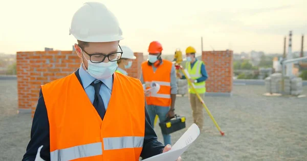 Portret kaukaskiego młodego biznesmena inwestora w hemet i maskę medyczną stojącego przy budynku z projektem budowlanym w rękach. Mężczyzna inżynier lub architekt na stronie konstrukcyjnej pandemii. — Zdjęcie stockowe