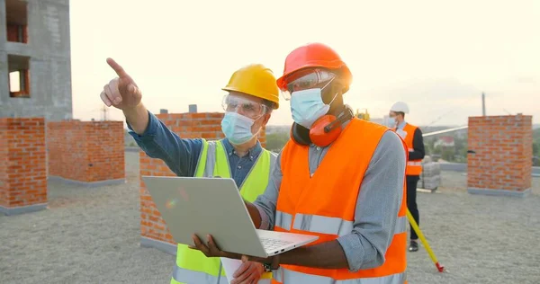 Grupo de construtores em hardhat trabalha no canteiro de obras. Dois construtores masculinos em máscara facial médica usando computador portátil no local de construção. — Fotografia de Stock