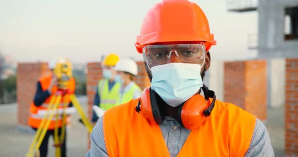 Retrato de construtor de homens afro-americanos ou gerente de construção em máscara médica e capacete de proteção com equipamentos de engenharia em pé no local de construção. — Fotografia de Stock