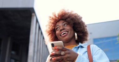 Afro-amerikan neşeli, stil sahibi genç bir bayanın telefon dinlemesi ya da kaydırması ve şehrin caddesinde durması..