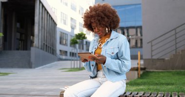 Afro-Amerikalı güzel bir kadın tablet üzerinde tıkırdıyor ve kaydırıyor, gülümsüyor ve dışarıda bankta oturuyor..