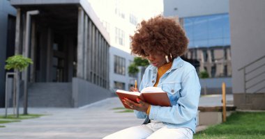 Genç kıvırcık ve güzel Afro-Amerikan kadın portresi bankta oturuyor, ders kitabı okuyor, notlar yapıyor ve kameraya gülümsüyor..