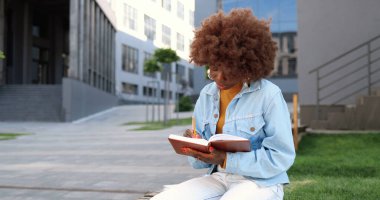 Genç kıvırcık ve güzel Afro-Amerikan kadın portresi bankta oturuyor, ders kitabı okuyor, notlar yapıyor ve kameraya gülümsüyor..