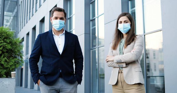 Портрет кавказского юноши и девушки в деловом стиле и в медицинских масках, стоящих на социальной дистанции и смотрящих в камеру. Бизнесмен и предпринимательница на открытом воздухе во время пандемии.