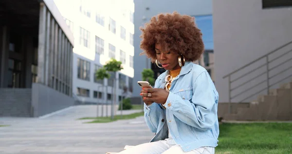 아프리카계 미국인아름다운 여성이 핸드폰으로 두드리고 스크롤하고 밖에서 벤치에 — 스톡 사진