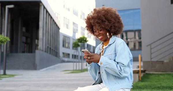 아프리카계 미국인아름다운 여성이 핸드폰으로 두드리고 스크롤하고 밖에서 벤치에 — 스톡 사진