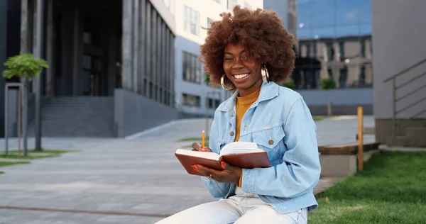 年轻的卷曲美丽的非洲裔美国女人坐在长椅上 阅读课本 在镜头前做记号和微笑的画像 — 图库照片