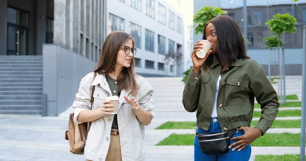 Mixed-races young pretty females, best friends talking cheerfully and walking with cups of coffee to-go and at city street.