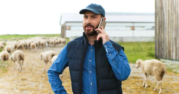 Красавчик улыбнулся кавказскому пастуху, разговаривающему по мобильному телефону, в то время как на заднем плане шла стая овец. Мужчина фермер говорит по телефону на улице в сарае. — стоковое фото