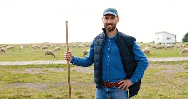 Retrato tiro de bonito branco jovem pastor em boné e com vara de pé ao ar livre no campo da fazenda. Ovelhas rebanho pastoreando em grama verde backgroung. Criação de gado. Exploração de lã. — Fotografia de Stock