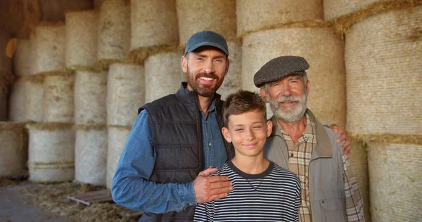 Três gerações masculinas em banhos de feno. avô caucasiano, pai e filho sorrindo para a câmera e em pé no celeiro. Conceito de agricultura. No campo. Velho agricultor com filho e neto. — Fotografia de Stock