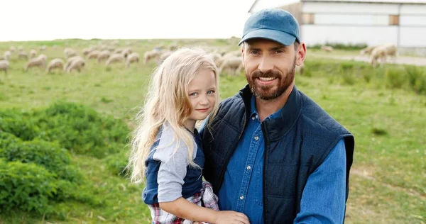 Portret van een blanke jonge knappe vader die zijn handen vasthoudt aan een schattig dochtertje en glimlacht naar de camera in het groene veld. Schapen grazen op de achtergrond een weiland. Glimlachte vrolijke man en klein meisje. — Stockfoto
