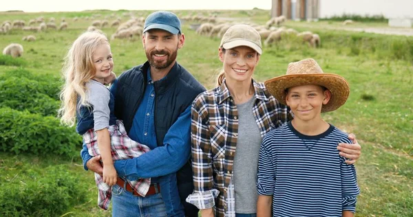 Retrato de família feliz caucasiana com crianças pequenas em pé no pasto com ovelhas rebanho no fundo e sorrindo para a câmera. Pais alegres com pequeno filho e filha no campo na fazenda. — Fotografia de Stock