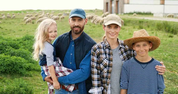 Retrato de família feliz caucasiana com crianças pequenas em pé no pasto com ovelhas rebanho no fundo e sorrindo para a câmera. Pais alegres com pequeno filho e filha no campo na fazenda. — Fotografia de Stock