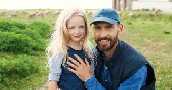 Portret van een blanke jonge knappe vader die buiten staat met een schattig dochtertje en glimlacht naar de camera in het groene veld. Schapen grazen op de achtergrond een weiland. Glimlachte man en klein meisje. — Stockfoto